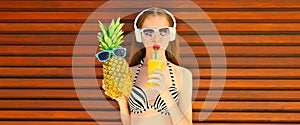 Summer portrait of happy young woman listening to music in headphones and drinking fresh juice holding pineapple in sunglasses