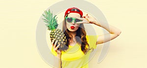 Summer portrait of happy young woman with pineapple blowing her lips with red lipstick wearing baseball cap, sunglasses on white