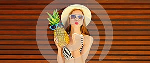 Summer portrait of happy young woman with pineapple blowing her lips sends air kiss wearing summer straw hat on wooden background