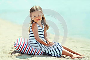 Summer portrait little girl child wearing striped dress sitting on a sand beach on a sea background