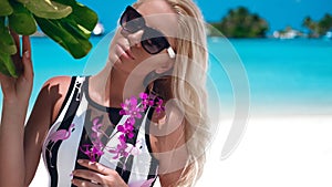 Summer portrait of Sexy blond Woman in fashionable sunglasses with purple flowers suntanning on tropical beach. Slim girl posing