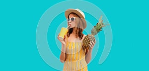 Summer portrait of stylish happy young woman drinks fresh juice with pineapple fruits wearing sunglasses, straw hat posing on blue