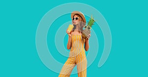 Summer portrait of young woman drinking fresh juice and holding pineapple wearing straw hat, sunglasses on blue background