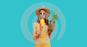 Summer portrait of young woman drinking fresh juice and holding pineapple wearing straw hat, sunglasses on blue background