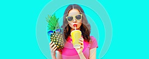 Summer portrait of young woman drinking juice and holding pineapple wearing sunglasses on blue background