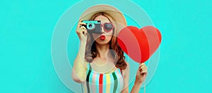 Summer portrait young woman taking a picture on retro camera with red heart shaped balloon wearing a straw hat on blue