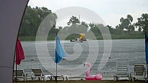 Summer rain, thunderstorm, heavy rainfall on beach, by river. lonely empty sun loungers and beach umbrellas are standing