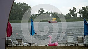 summer rain, thunderstorm, heavy rainfall on beach, by river. lonely empty sun loungers and beach umbrellas are standing