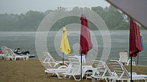 Summer rain, thunderstorm, heavy rainfall on an empty beach, by river. lonely empty sun loungers and beach umbrellas are