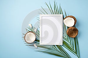 Summer rest concept. Top view photo of white photo frame cracked coconuts and green palm leaves on isolated light blue background