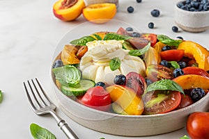 Summer salad with burrata cheese, grilled peaches, tomatoes, blueberries, cucumber, olive oil and basil in a plate