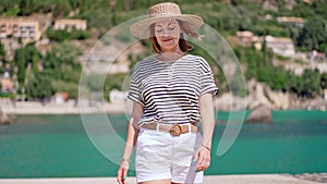 Summer scene - smiling woman in straw hat walking to camera. Sea, motor boat on background. Girl on pier in Greece or