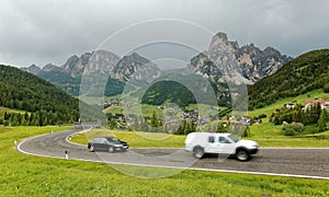 Summer scenery of Dolomiti with villages on the grassy hillside of rugged mountains & cars traveling on a highway