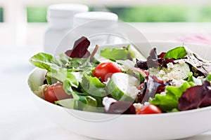 Summer season salad with salad leaves, tomatoes, cucumbers, Italian herbs and cheese in a bowl on a table