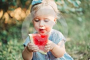 Summer seasonal picnic food. Cute baby girl eating ripe red watermelon in park. Funny child kid sitting on ground with fresh