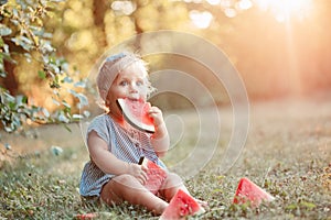 Summer seasonal picnic food. Cute Caucasian baby girl eating ripe red watermelon in park. Funny child kid sitting on ground with