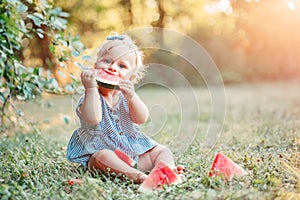 Summer seasonal picnic food. Cute Caucasian baby girl eating ripe red watermelon in park. Funny child kid sitting on ground with