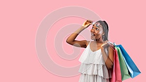 Summer shopping. Happy black woman with bags lifting sunglasses, looking at empty space on pink background