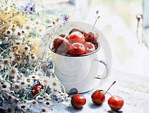 Summer simple still life. Sweet cherry in an iron mug and a bouquet of field tender flowers on the old window overlooking the gard