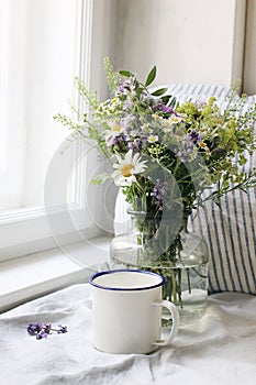 Summer still life scene. Enamel mug, wild flowers bouquet and linen cushion. Vintage feminine photo, rustic design