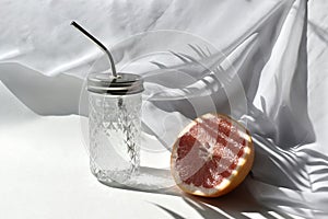 Summer still life vacation scene. Close up fresh water in cocktail glass and cut grapefruit on white table in sunlight.