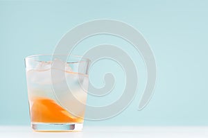 Summer striped oranges lemonade with ice cube, liquor in misted glass on elegant pastel green wall, white wood table.