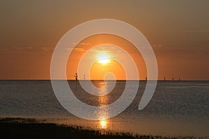 The summer sun sets amid a brilliant orange sky at a calm ocean harbor.