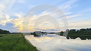 The summer sun sinks behind the trees growing on the riverbank. The colorful cloudy sky with feathery clouds at sunset is reflecte