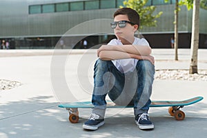 Summer sunny day. Boy in white T-shirt and sunglasses sitting outdoor on longboard. Boy rests after skiing on longboard