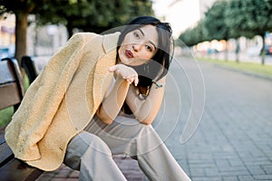 Summer sunny lifestyle fashion portrait of young stylish Asian woman, wearing trendy colorful outfit, sitting on city
