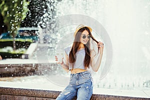 Summer sunny lifestyle fashion portrait of young stylish hipster woman walking on the street, wearing cute trendy outfit, drinking