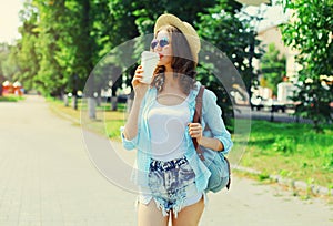 Summer sunny portrait beautiful smiling young woman with cup of coffee wearing a straw hat and backpack in the city park