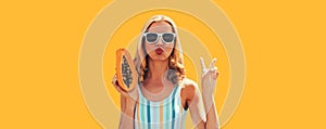 Summer sunny portrait of stylish young woman with fresh juicy papaya fruit blowing her lips sending air kiss on orange background