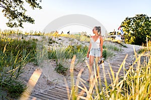 Summer sunset walk on beach boardwalk, green grass field and sand dunes. Outdoor nature portrait of happy pretty girl.