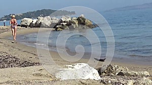 Summer time.  Young woman in swimsuit runs from right to left on water on beach. View from the back. Beautiful place with rocks