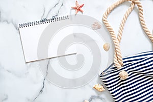 Summer travel composition on a marble background. Women`s desk with striped beach bag, seashells and blank notepad. Lifestyle of