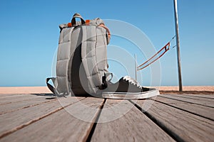 Summer travel concept. Backpack and sneakers are on the wooden desk near the beach and sea.Blue clear sky and sun.