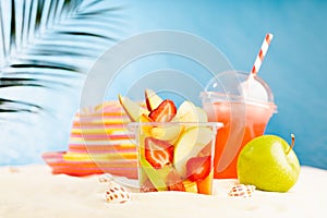 Summer travel and holiday background - picnic with cold drink, fresh fruit salad takeaway, sun hat on hot sunny sandy beach.