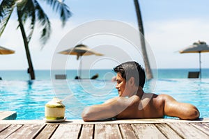 Summer travel vacation concept, Traveler asian man with coconut relax in luxury infinity pool hotel resort with sea beach and palm