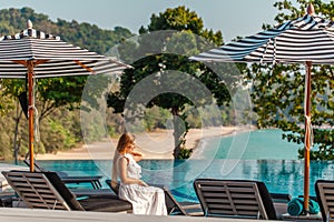 Traveler woman in white dress and hat sitting on sun lounger near swimming pool