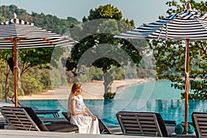 Traveler woman in white dress and hat sitting on sun lounger near swimming pool