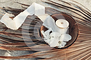 Summer tropical lifestyle scene. Oleander blossoms and white silk ribbons spools in coconut shell. Dry palm leaves