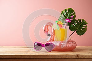 Summer tropical vacation concept with orange juice and flamingo pool float on wooden table over pink background