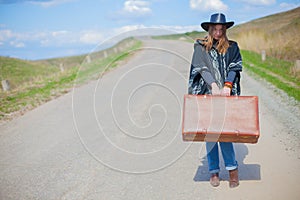 A girl in blue jeans, a poncho, a black leather hat with an old brown suitcase in her hands is on the road.