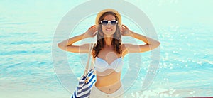 Summer vacation, beautiful happy smiling woman in bikini swimsuit and straw hat with bag on the beach on sea coast background on