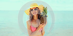 Summer vacation, beautiful happy smiling woman in bikini swimsuit and straw hat holding pineapple fruits on the beach on sea coast