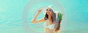 Summer vacation, beautiful happy smiling woman in bikini swimsuit and straw hat holding pineapple fruits on the beach on sea coast
