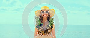 Summer vacation, beautiful happy smiling woman in bikini swimsuit and straw hat holding pineapple fruits on the beach on sea coast