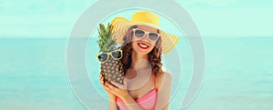 Summer vacation, beautiful happy smiling woman in bikini swimsuit and straw hat holding pineapple fruits on the beach on sea coast