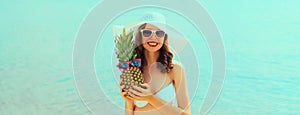 Summer vacation, beautiful happy smiling woman in bikini swimsuit and straw hat holding pineapple fruits on the beach on sea coast